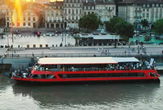 Bateau de croisière rouge et blanc naviguant sur la Garonne à Bordeaux, avec des bâtiments historiques en arrière-plan et le soleil couchant illuminant la skyline de la ville.