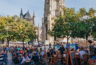 Vue d'une terrasse animée sur une place de Bordeaux avec la basilique Saint-Michel en arrière-plan, sous un ciel bleu dégagé.