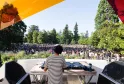 Un DJ en chemise rayée joue sur scène devant une foule importante dans un parc de Bordeaux, avec des parasols colorés et des arbres en arrière-plan.