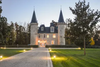 Château avec deux tours pointues illuminées la nuit, entouré d'arbres et d'une allée pavée éclairée, près de Bordeaux