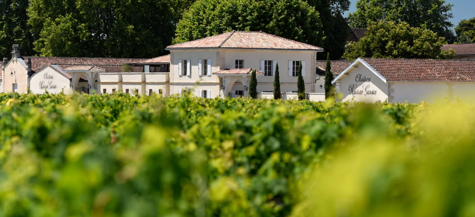 Château viticole bordelais avec façade blanche et toits en tuiles, entouré de vignobles verdoyants et de forêts.