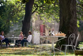 Table dressée en plein air sous les arbres avec verres, bouteilles de vin et pain, tandis que des personnes dégustent à l'arrière-plan dans un jardin bordelais.