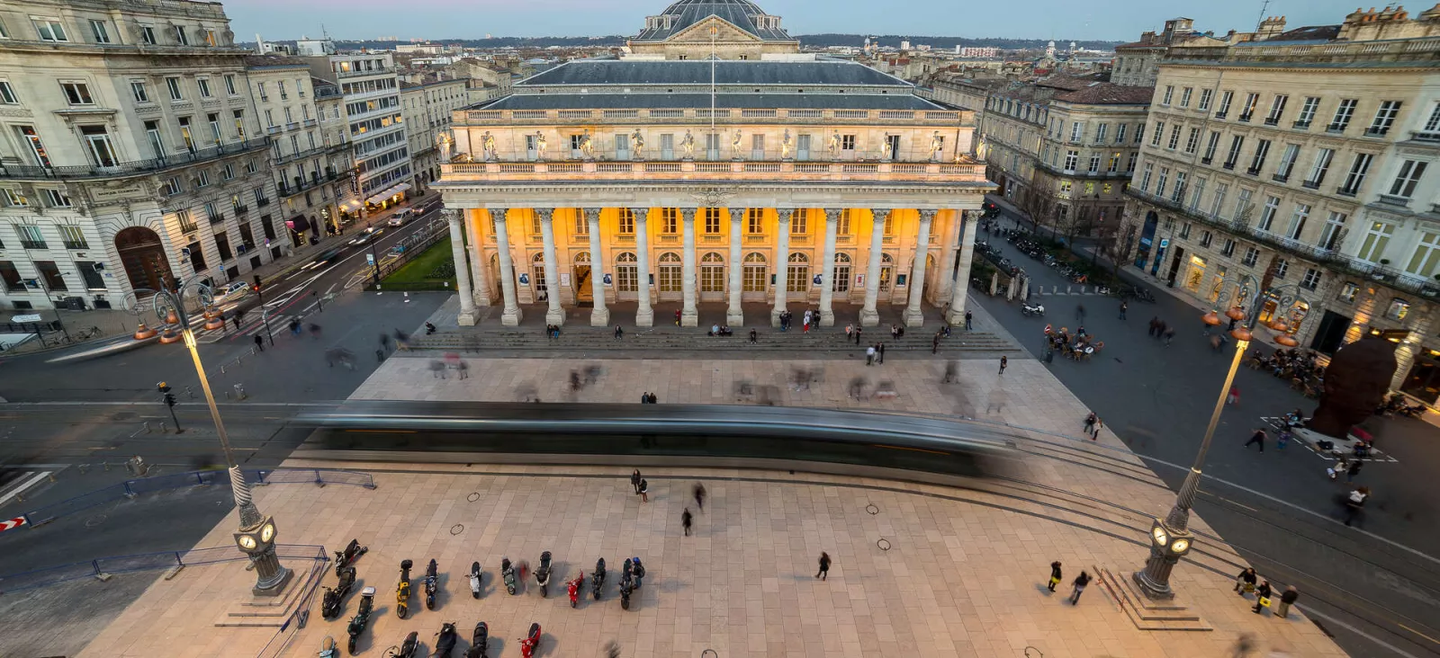 Vue aérienne de la Place de la Comédie à Bordeaux avec le Grand-Théâtre illuminé au centre, entouré de bâtiments haussmanniens et de personnes se promenant sur la place.