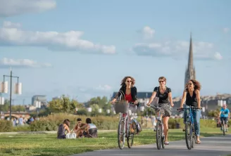 Trois femmes à vélo sur une piste cyclable dans un parc de Bordeaux, avec la basilique Saint-Michel visible en arrière-plan et des promeneurs sur l'herbe.