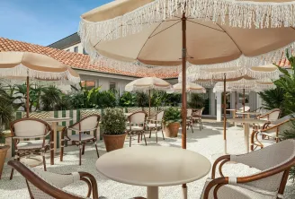 Terrasse élégante d'un hôtel bordelais avec parasols écru à franges, mobilier en rotin, plantes en pots et vue sur toiture en tuiles romaines
