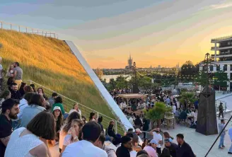 Vue panoramique d'une zone urbaine de Bordeaux avec une terrasse en pente couverte de végétation, des visiteurs profitant du coucher de soleil, et la skyline de la ville avec ses églises historiques en arrière-plan.